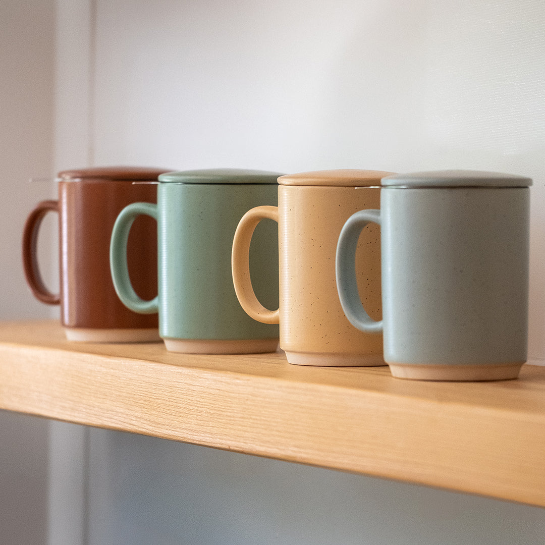 Four ceramic mugs with lids and metal tea straws lined up on a light wood shelf against a neutral background.