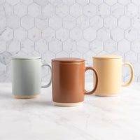 Three ceramic lidded mugs with speckled glaze in mocha mousse, sand, and sky, arranged on a white marble surface against a white hexagonal tile backsplash.