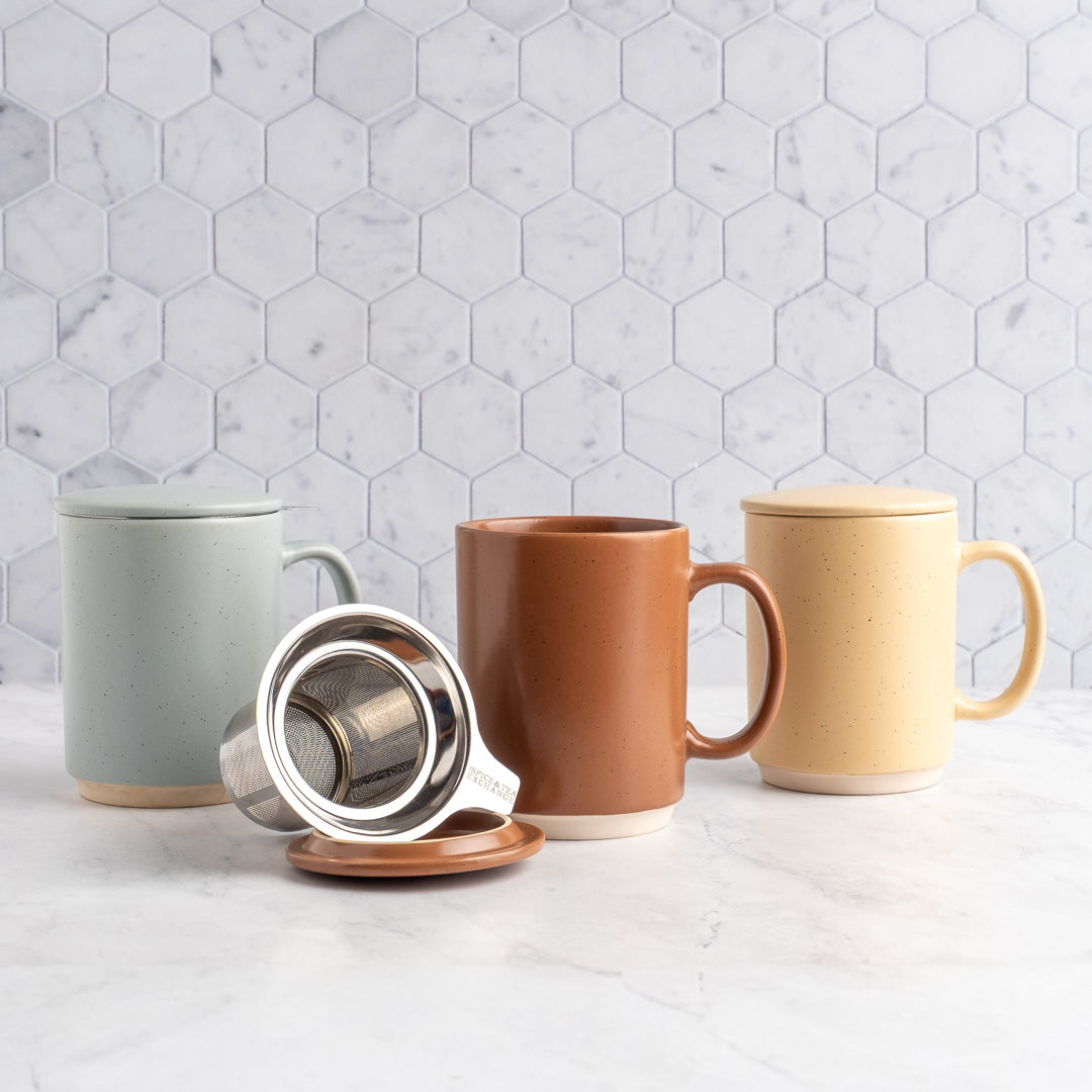 Three ceramic mugs with lids in mocha mousse, sand, and sky arranged on a white marble surface with a white hexagonal tile backsplash. A stainless steel tea infuser and terracotta lid rest in front of the mugs.