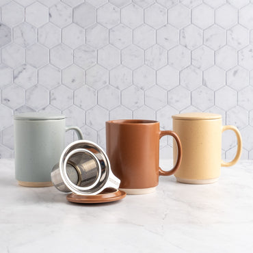 Three ceramic mugs with lids in mocha mousse, sand, and sky arranged on a white marble surface with a white hexagonal tile backsplash. A stainless steel tea infuser and terracotta lid rest in front of the mugs.