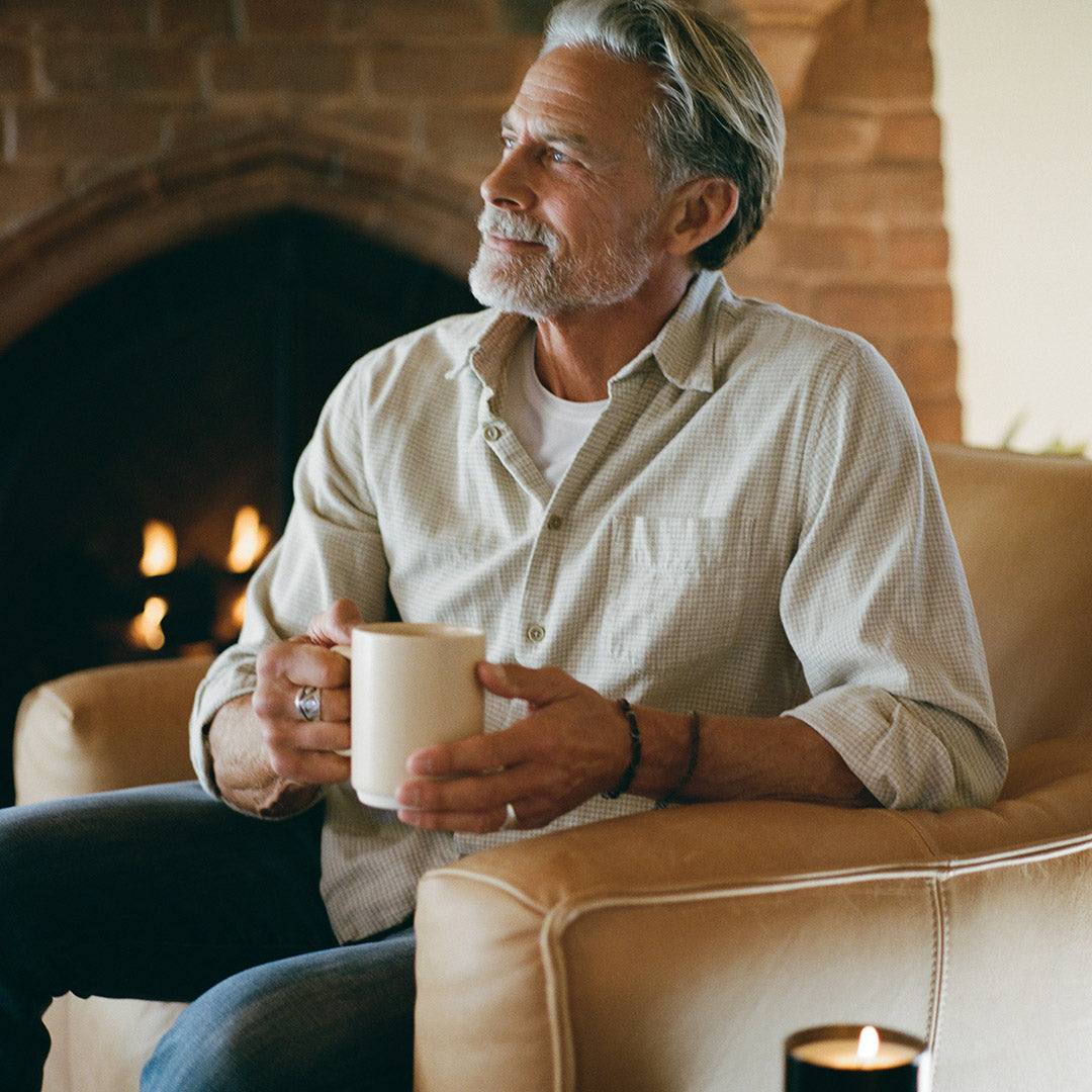 A man with gray hair and a beard sits in a beige armchair in front of a brick fireplace, holding a sand colored ceramic mug. He is wearing a light beige button-down shirt over a white t-shirt and blue jeans.