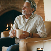 A man with gray hair and a beard sits in a beige armchair in front of a brick fireplace, holding a sand colored ceramic mug. He is wearing a light beige button-down shirt over a white t-shirt and blue jeans.