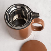 A mocha mousse ceramic mug with a stainless steel filter insert and a matching lid, shown on a light marble surface. The handle is engraved with 'SPICE&TEA EXCHANGE'.