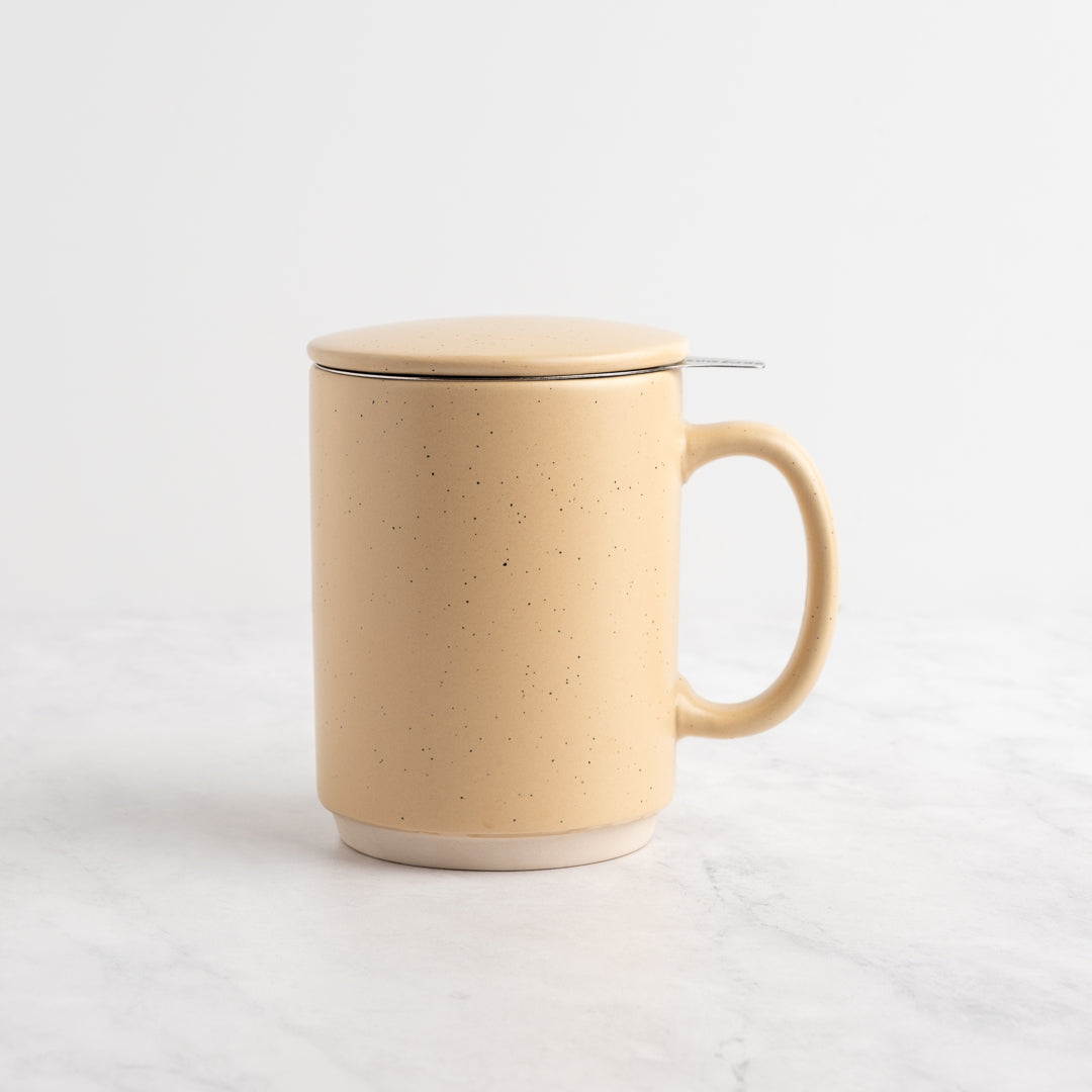 A sand speckled ceramic mug with a matching lid and a small metal tea infuser, set on a white marble surface against a white background.