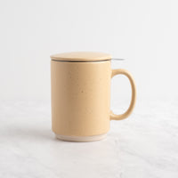 A sand speckled ceramic mug with a matching lid and a small metal tea infuser, set on a white marble surface against a white background.