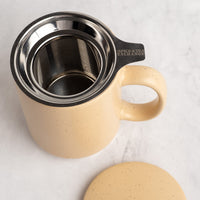 A sand speckled ceramic mug with an integrated stainless steel filter and a black handle, shown on a light marble surface with its matching lid placed nearby. The handle is marked 'SPICE&TEA EXCHANGE'.