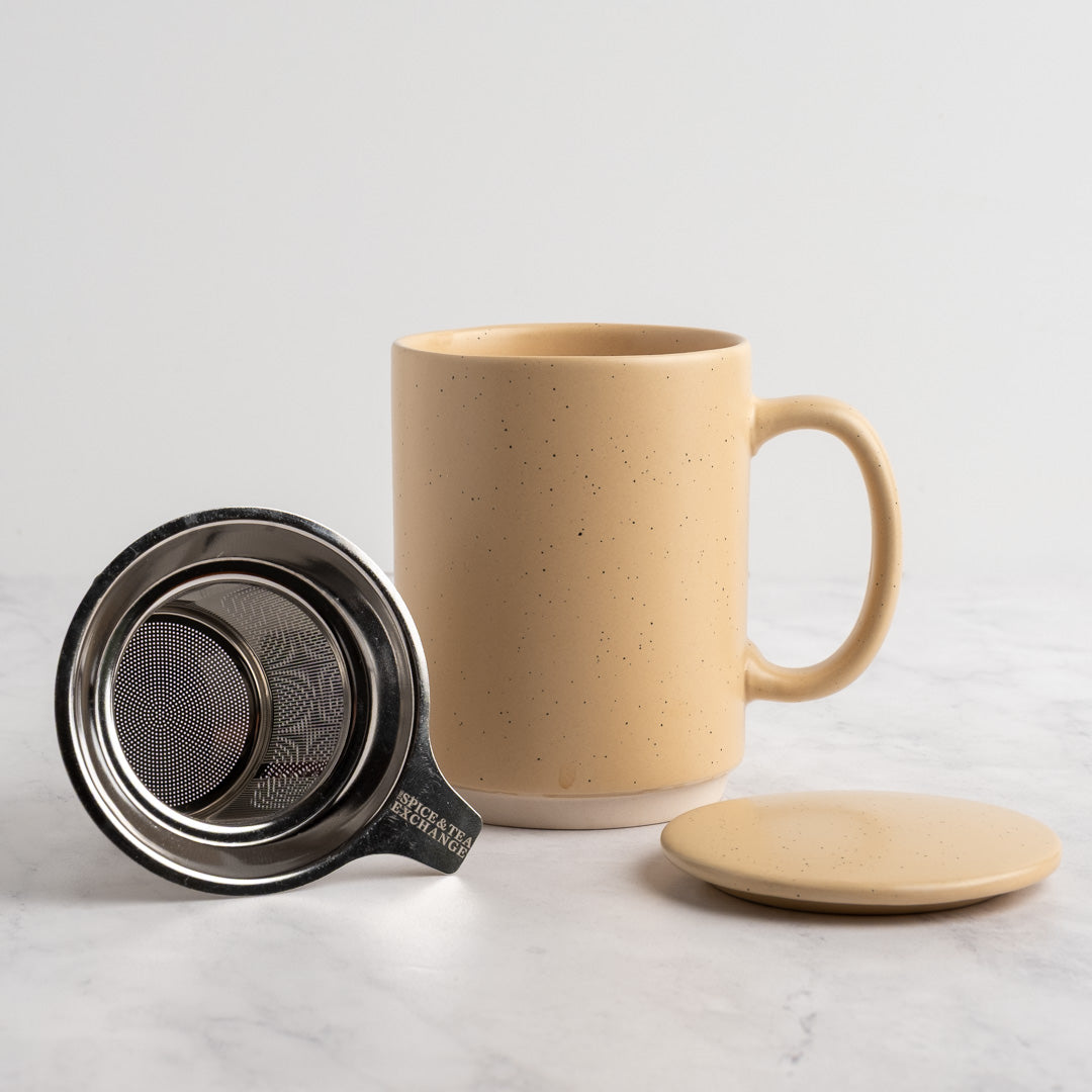 A speckled sand ceramic mug with a matching lid and metal tea infuser, displayed on a light marble surface against a white background.