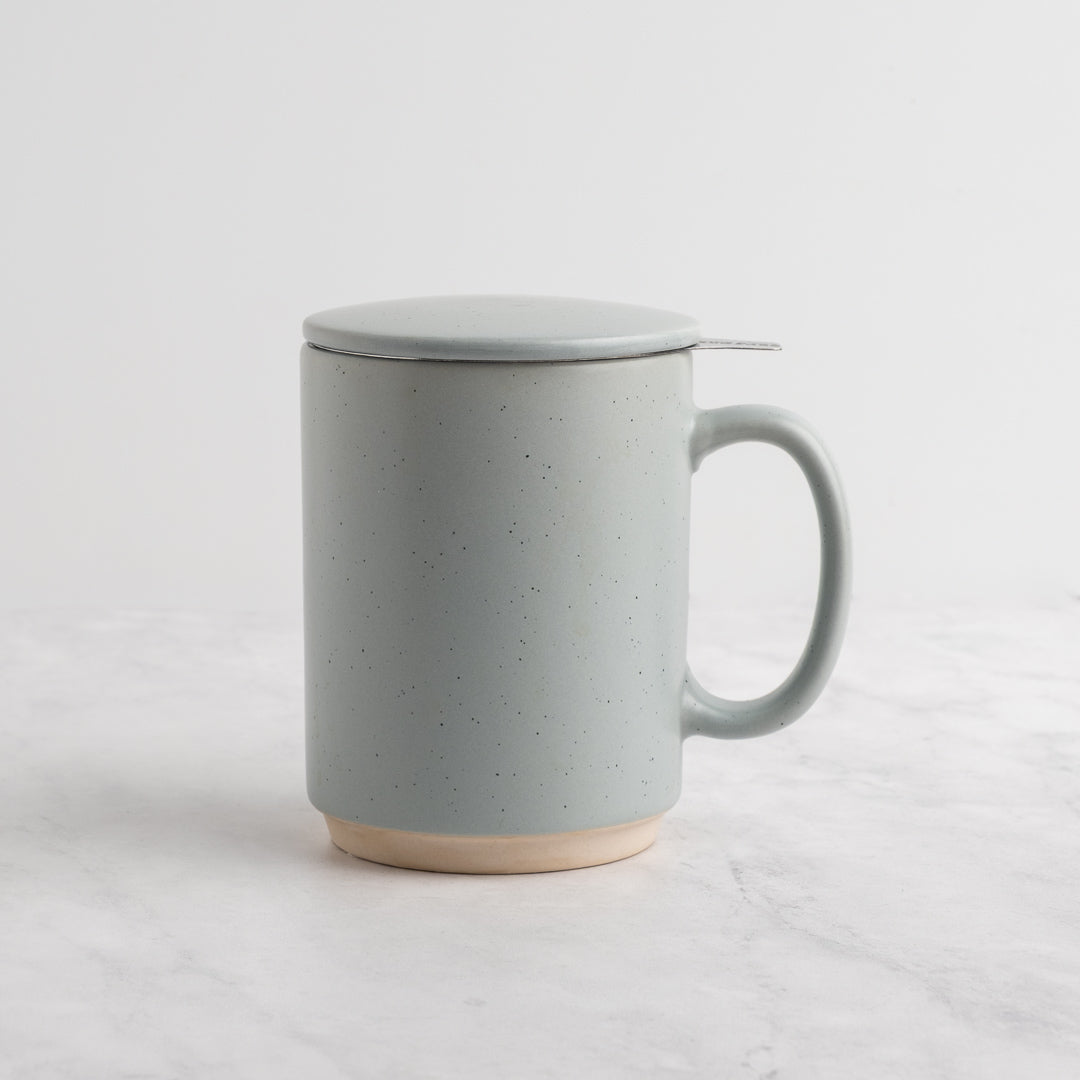 A speckled light blue-gray (sky) ceramic mug with a matching lid and a small metal spoon resting on top, set on a light marble surface against a white background.