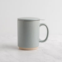A speckled light blue-gray (sky) ceramic mug with a matching lid and a small metal spoon resting on top, set on a light marble surface against a white background.