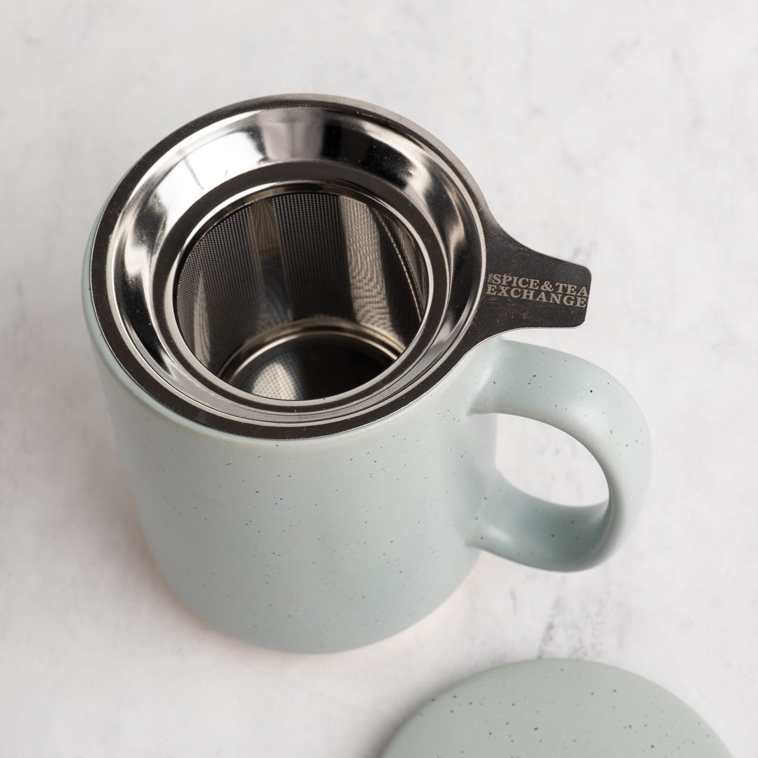 A speckled light blue gray (sky) ceramic mug with a stainless steel tea infuser inside, shown on a light marble surface with its matching lid partially visible in the foreground. The handle is engraved with 'SPICE & TEA EXCHANGE'.
