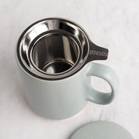 A speckled light blue gray (sky) ceramic mug with a stainless steel tea infuser inside, shown on a light marble surface with its matching lid partially visible in the foreground. The handle is engraved with 'SPICE & TEA EXCHANGE'.