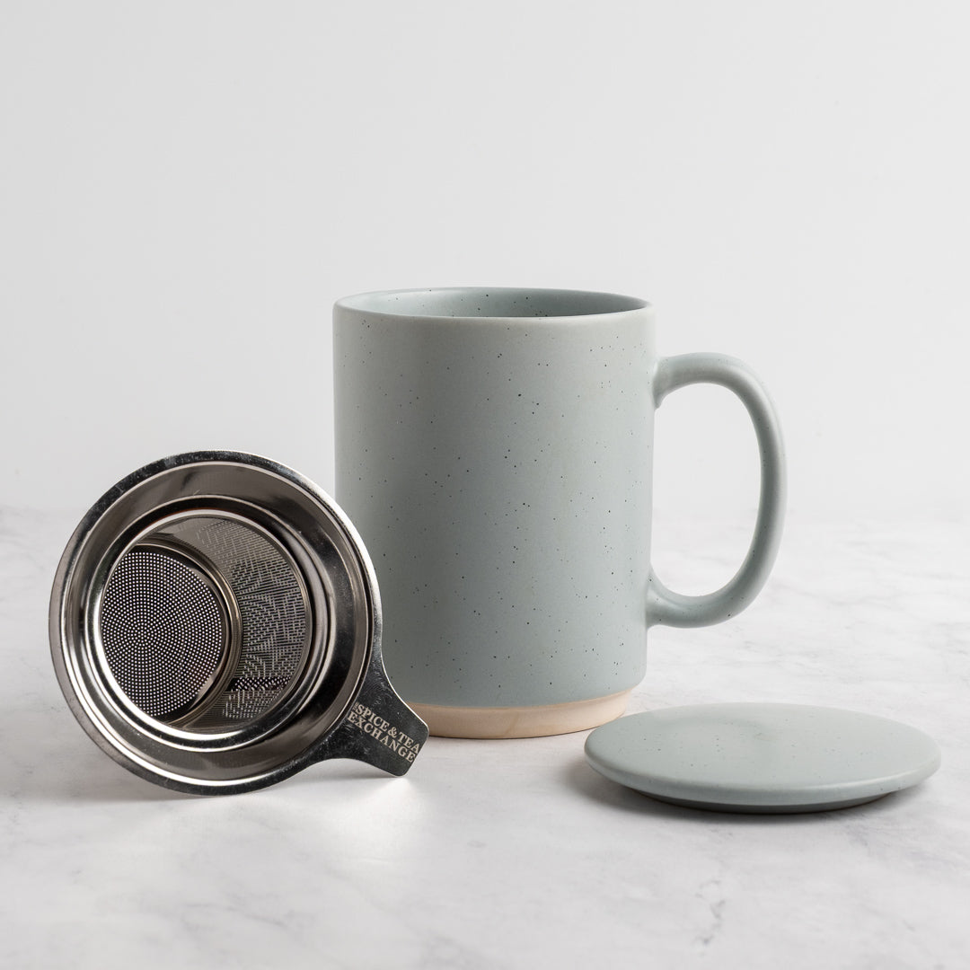 A speckled light blue-gray (sky) ceramic mug with a matching lid and stainless steel tea infuser, displayed on a light marble surface against a white background.