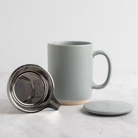 A speckled light blue-gray (sky) ceramic mug with a matching lid and stainless steel tea infuser, displayed on a light marble surface against a white background.