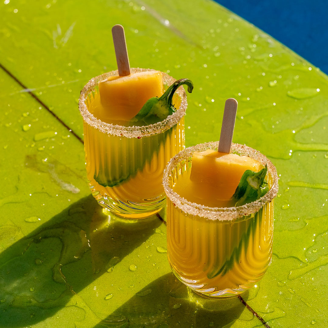 Two glasses of yellow cocktail with salted rims sit on a wet green surface. Each drink contains a mango popsicle and a green chili garnish, with sunlight casting bold shadows.