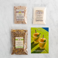 Four packaged tea and both product bags arranged on a light marble surface, with a printed card reading 'Chili Mango Poptail' at the bottom right.