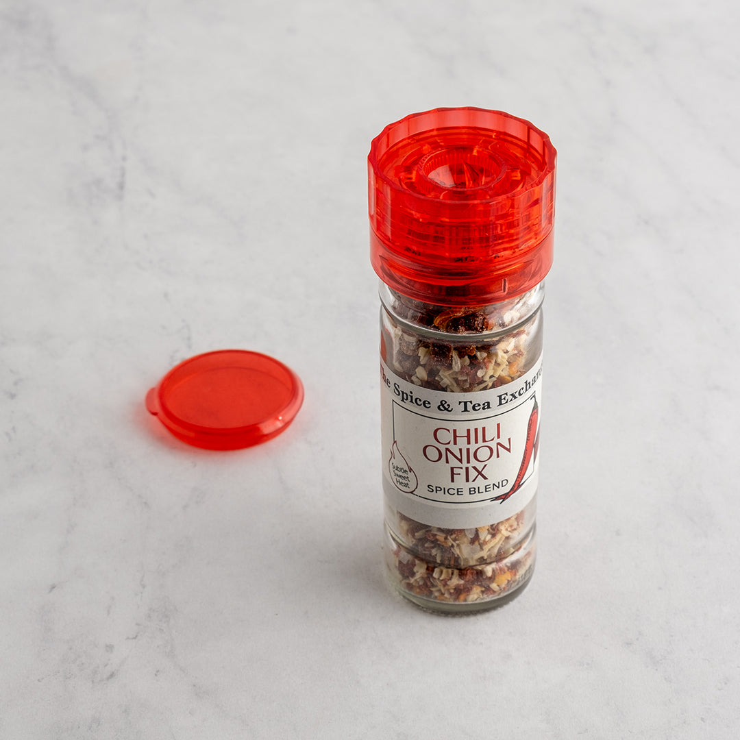 A clear glass spice jar labeled 'Chili Onion Fix' with a translucent red plastic lid, shown on a light marble surface with the red lid placed beside it.