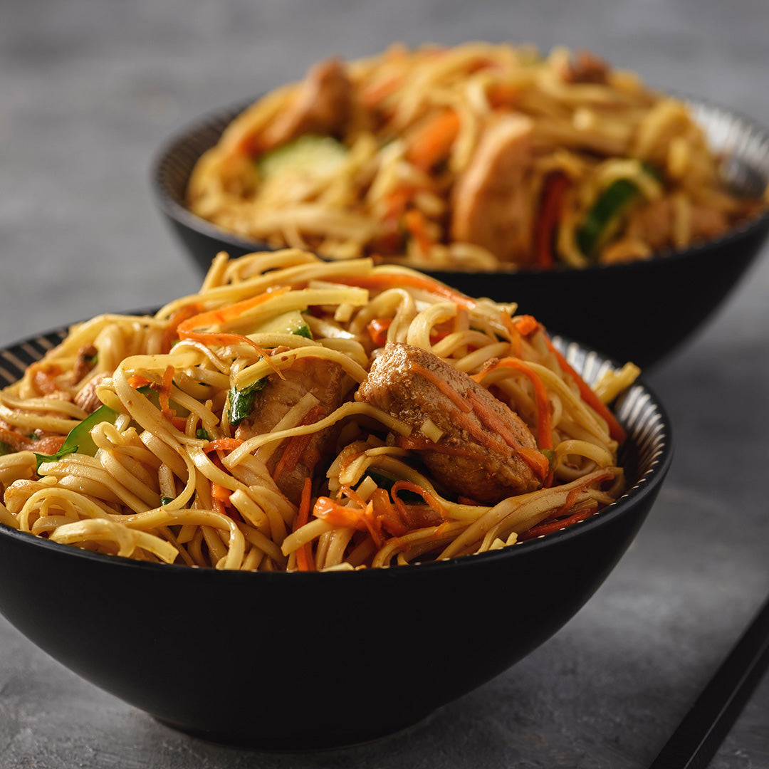 Two black bowls filled with stir-fried noodles, carrots, vegetables, and pieces of chicken, photographed on a gray surface with shallow depth of field.