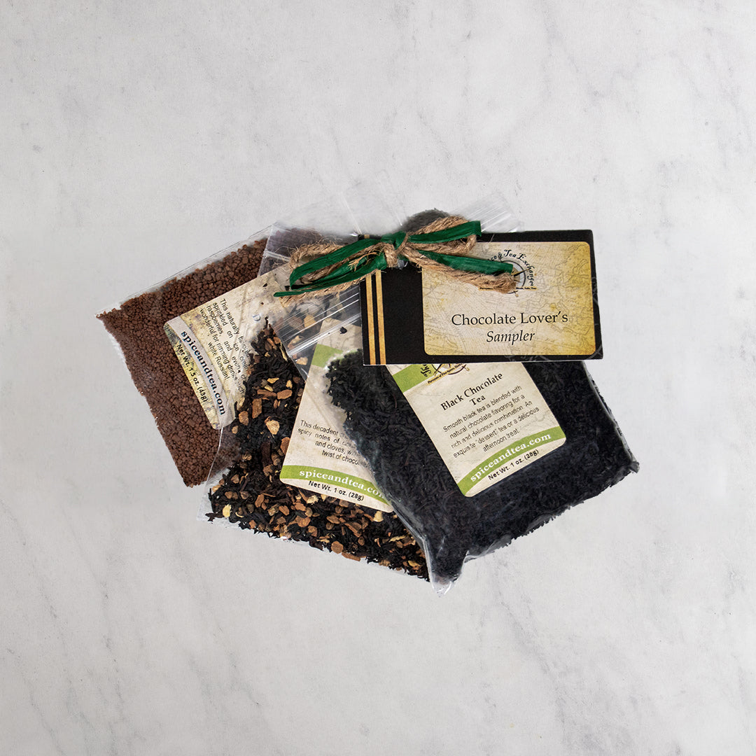 A packaged chocolate sampler gift set tied with a green ribbon, placed on a light marble surface. The set includes three small bags of tea, with a card reading 'Chocolate Lover's Sampler'.