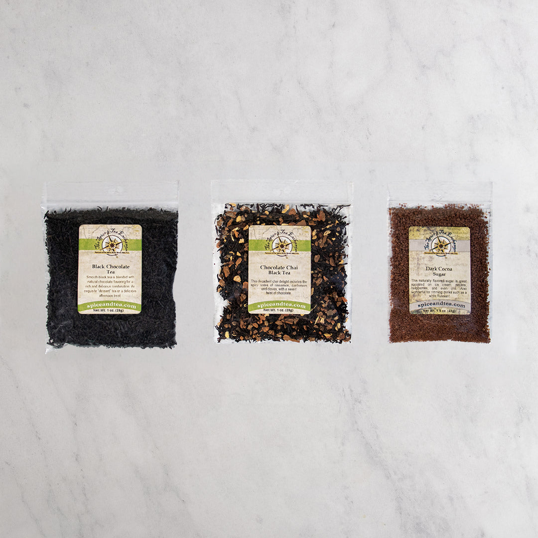 Three clear plastic bags of chocolate tea and a sugar arranged side by side on a white marble surface.