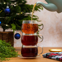 Three stacked clear glass teacups filled with tea, with hot tea being poured from a white teapot. The cups sit on a wooden table with a Christmas tree, greenery, and a plaid cloth in the background.