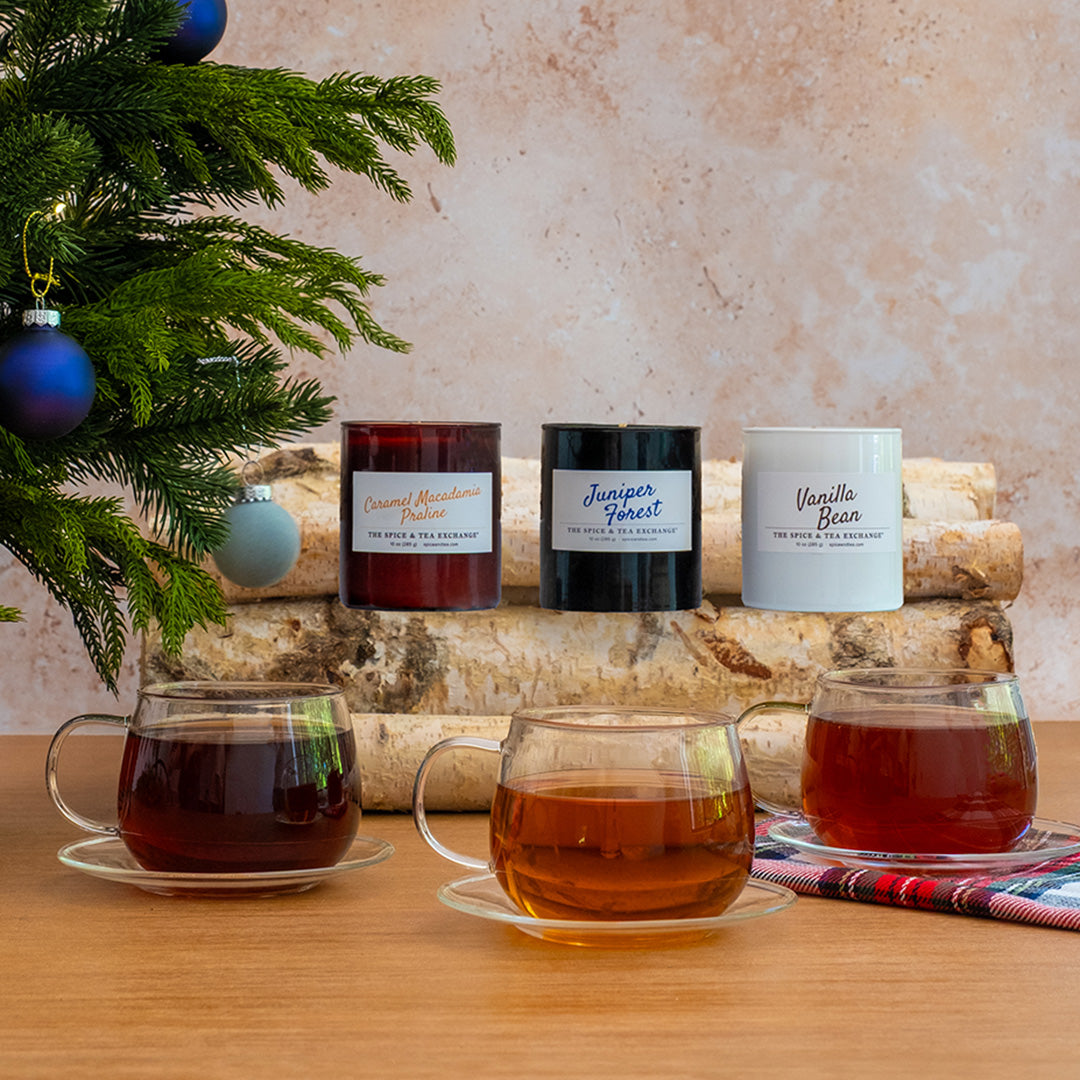 Three scented candles in glass jars are displayed on a birch log with three matching glass teacups filled with tea in the foreground.