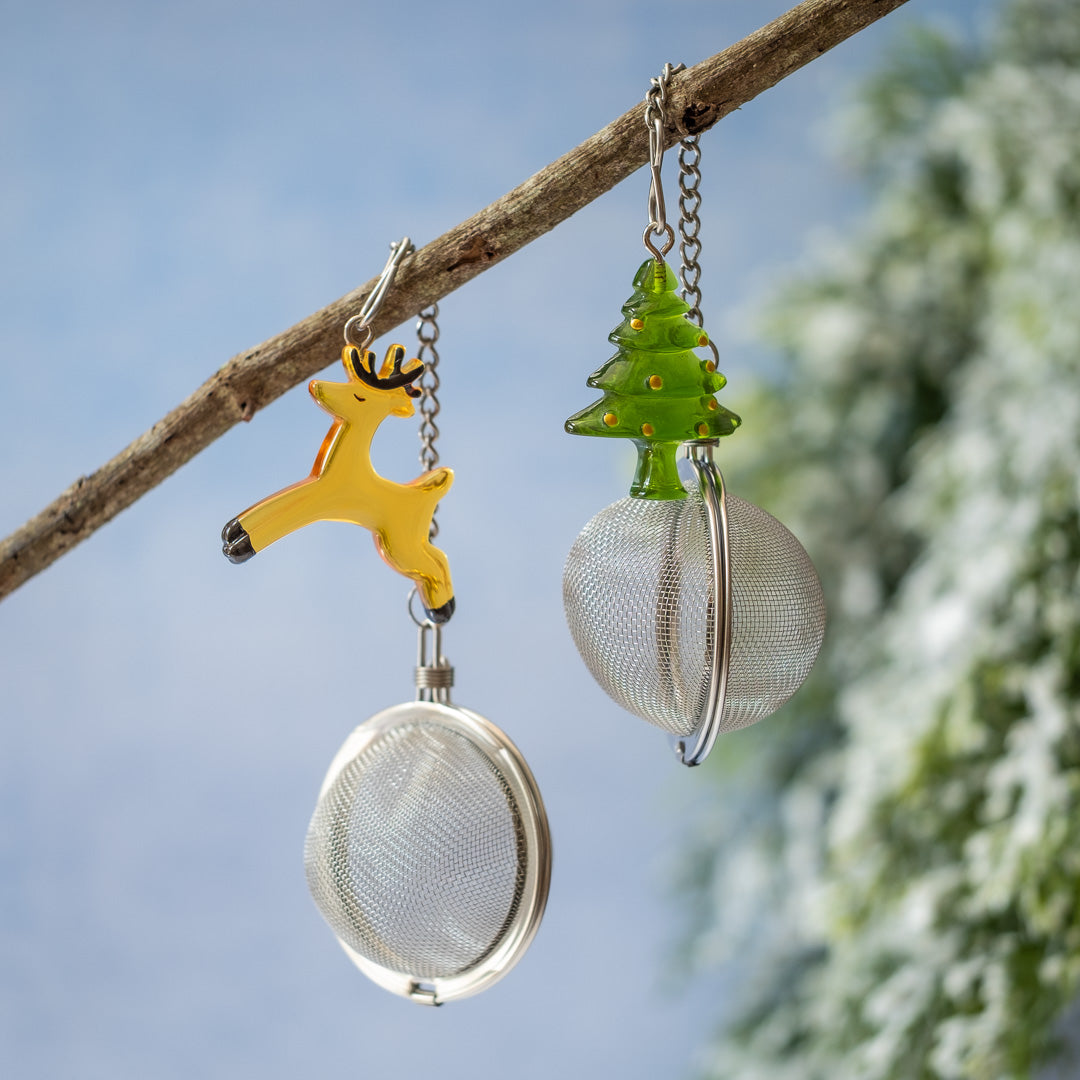 Two decorative tea infusers shaped like reindeer and a Christmas tree, hanging from a branch against a blue sky.