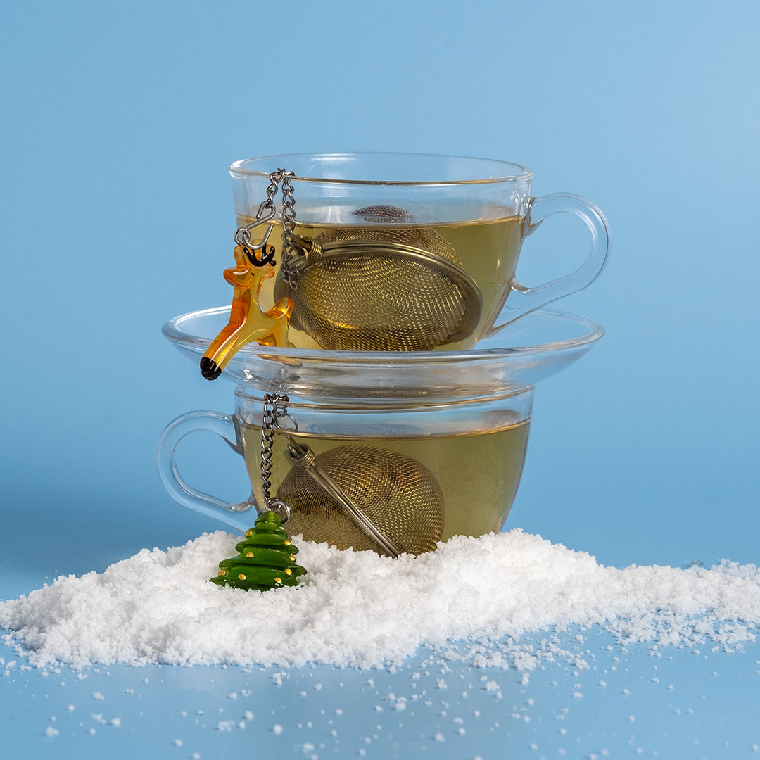Two stacked clear glass teacups filled with pale yellow tea, each holding a metal tea infuser and decorated with a hanging ornament shaped like a Reindeer and a green Christmas tree, set on white snow against a light blue background.