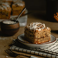A square slice of Cinnamon Vanilla Coffee Cake (Kit) with white icing drizzle rests on a small plate atop a striped cloth, accompanied by a fork and blurred baking ingredients in the background.