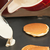 White pancake batter being poured onto a black griddle, with two golden-brown pancakes cooking on the surface and a red mixing bowl visible in the background.