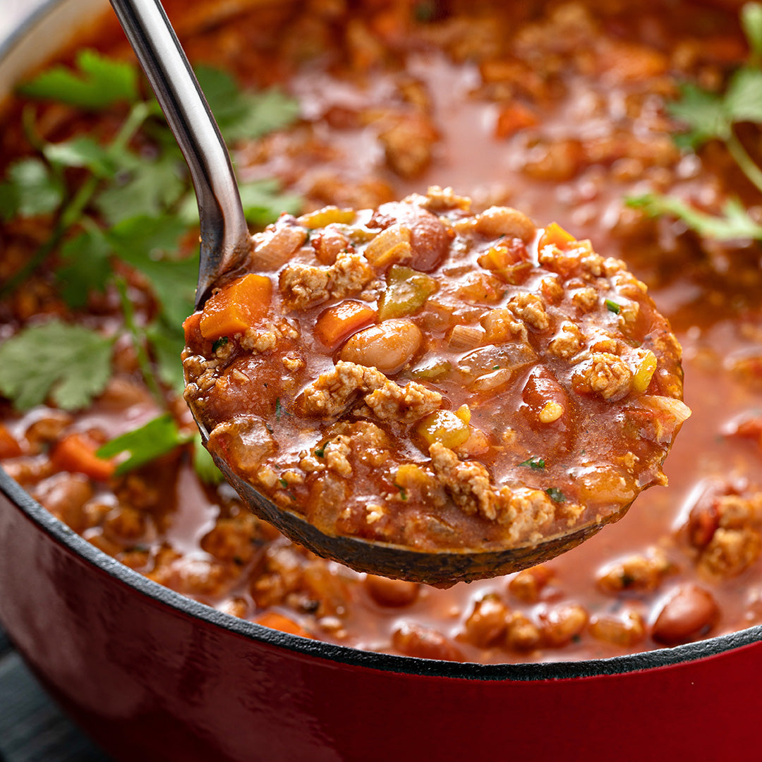 A close-up of a ladle filled with chunky meat and bean chili, held above a red pot of soup with fresh cilantro garnish.