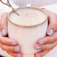 Person holding a mug of frothy beverage with a spoon, against a neutral background