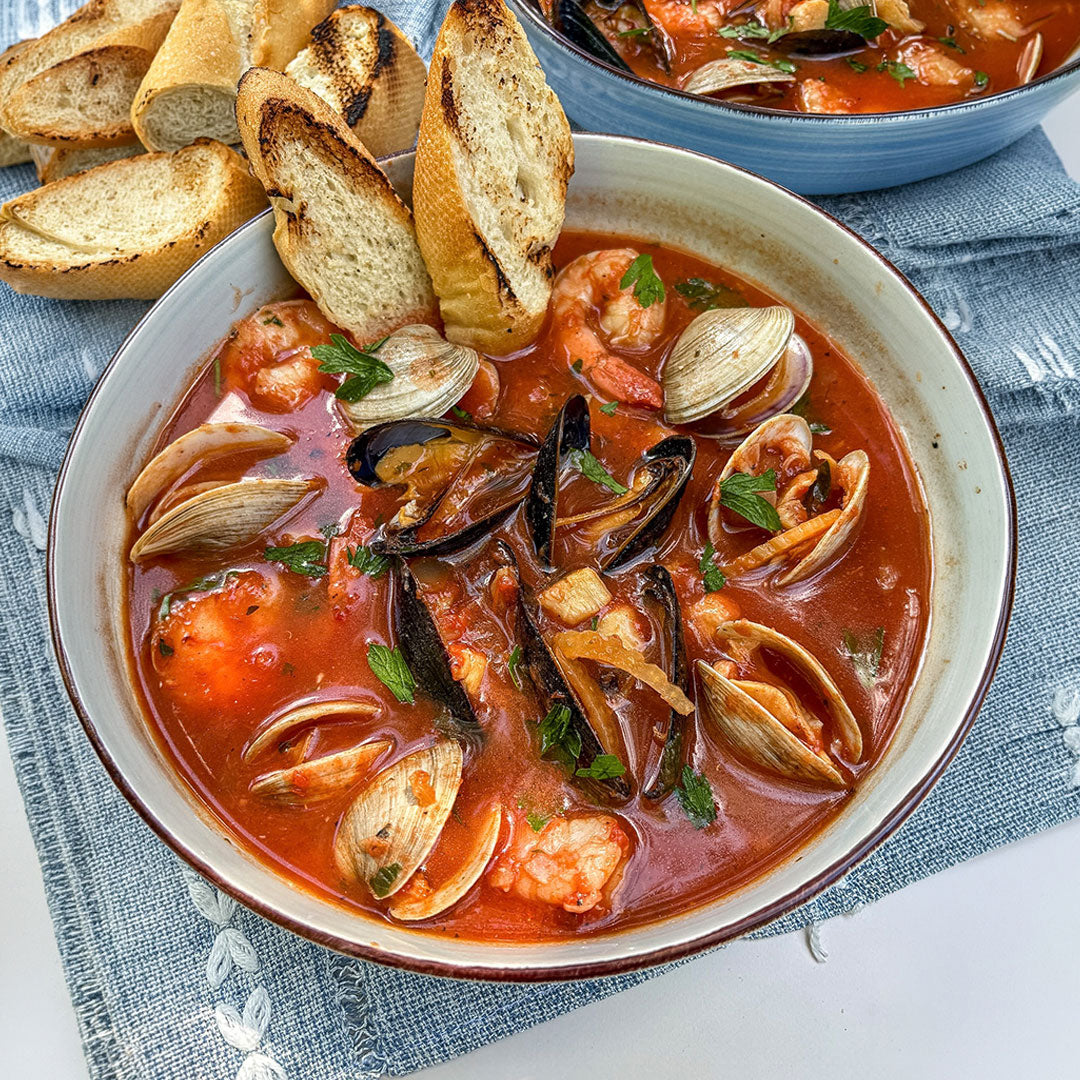 A close-up overhead shot of a bowl of seafood, featuring clams, mussels, and shrimp in a red-orange tomato-based sauce, served with toasted baguette slices on a blue textured cloth.