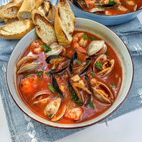 A close-up overhead shot of a bowl of seafood, featuring clams, mussels, and shrimp in a red-orange tomato-based sauce, served with toasted baguette slices on a blue textured cloth.