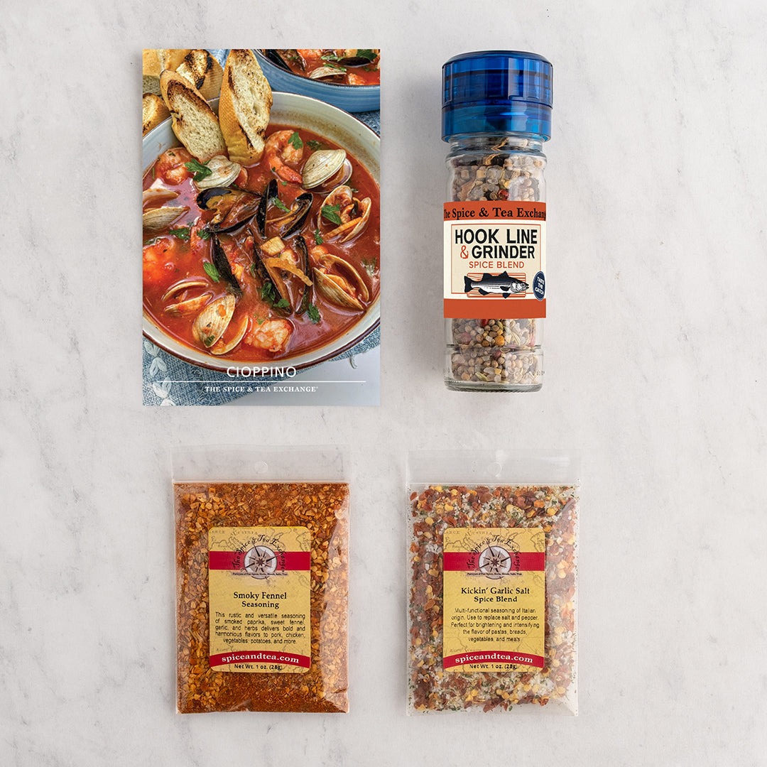 Cioppino (Seafood Stew) Recipe Card next to a glass spice grinder labeled 'Hook Line & Grinder', with two seasoning packets below on a white marble surface.