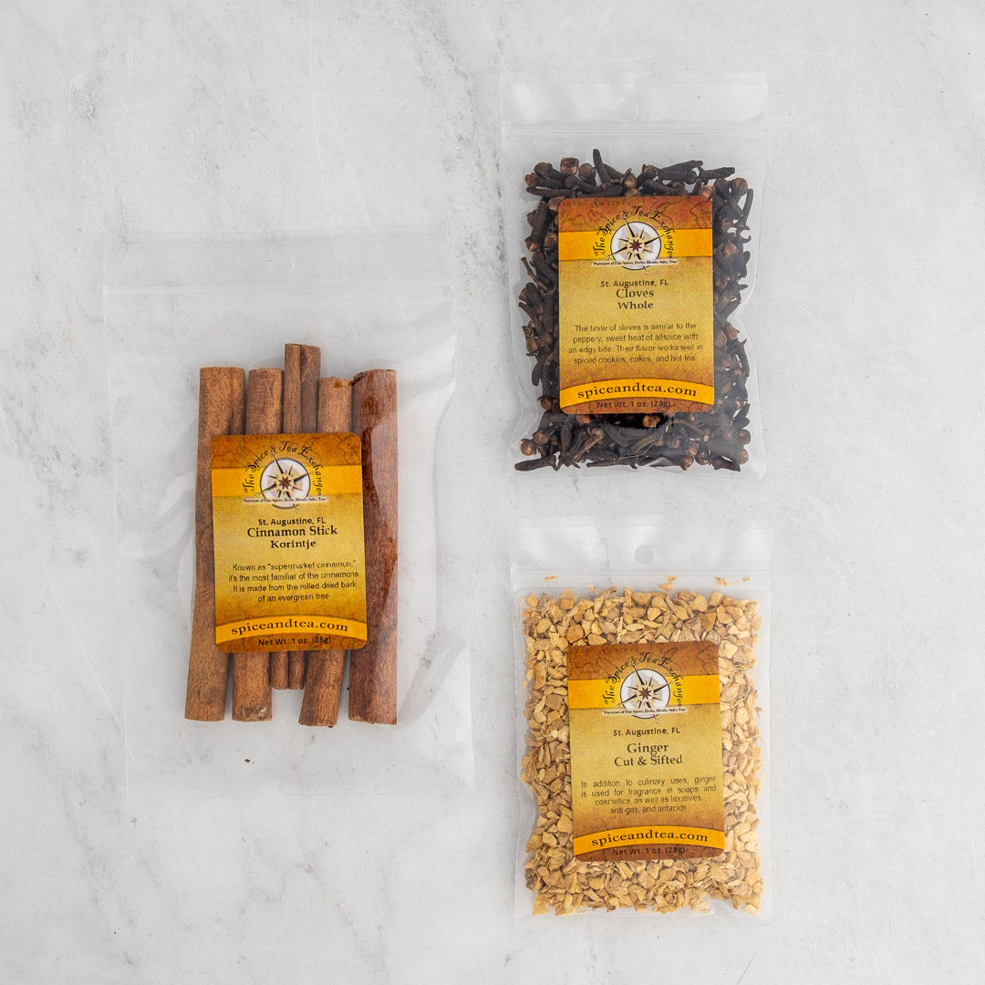 The Classic Cozy Simmer Pot Sampler features three clear bags with yellow labels—filled with dried cinnamon sticks, whole cloves, and cut ginger—perfect for filling your home with inviting fragrance. Displayed on a light marble surface.