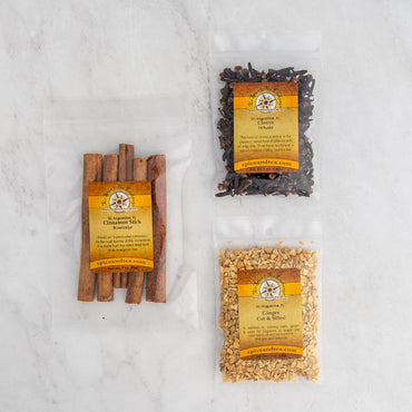 The Classic Cozy Simmer Pot Sampler features three clear bags with yellow labels—filled with dried cinnamon sticks, whole cloves, and cut ginger—perfect for filling your home with inviting fragrance. Displayed on a light marble surface.