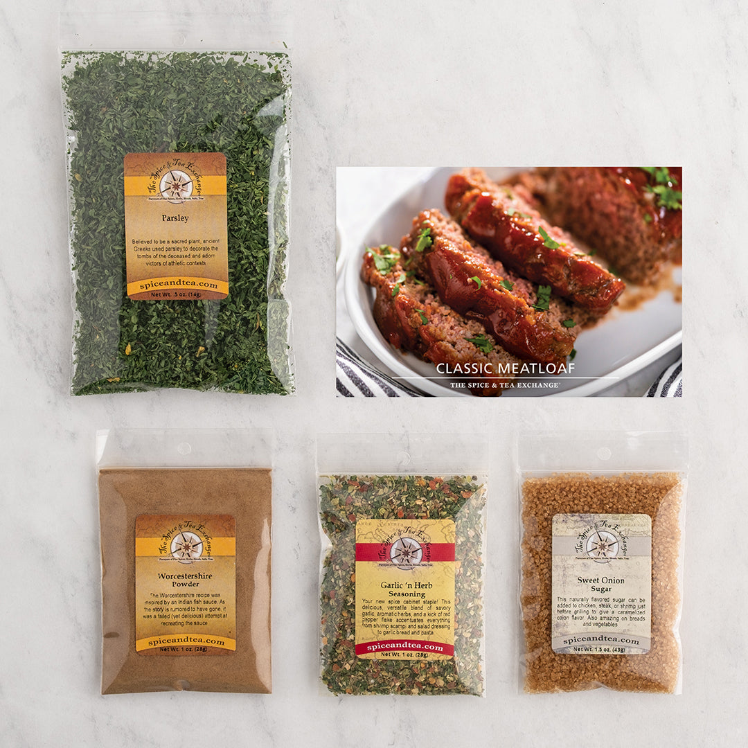 A flat-lay product arrangement on a white marble surface featuring: a large bag of parsley, a package of garlic and herb seasoning, a package of sweet onion sugar, a package of Worcestershire powder and a recipe card showing glazed classic meatloaf slices on a plate.