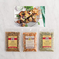 Three packaged seasoning blends are displayed on a white marble surface below a photo of grilled chicken kabobs with herbs and a side of dipping sauce.