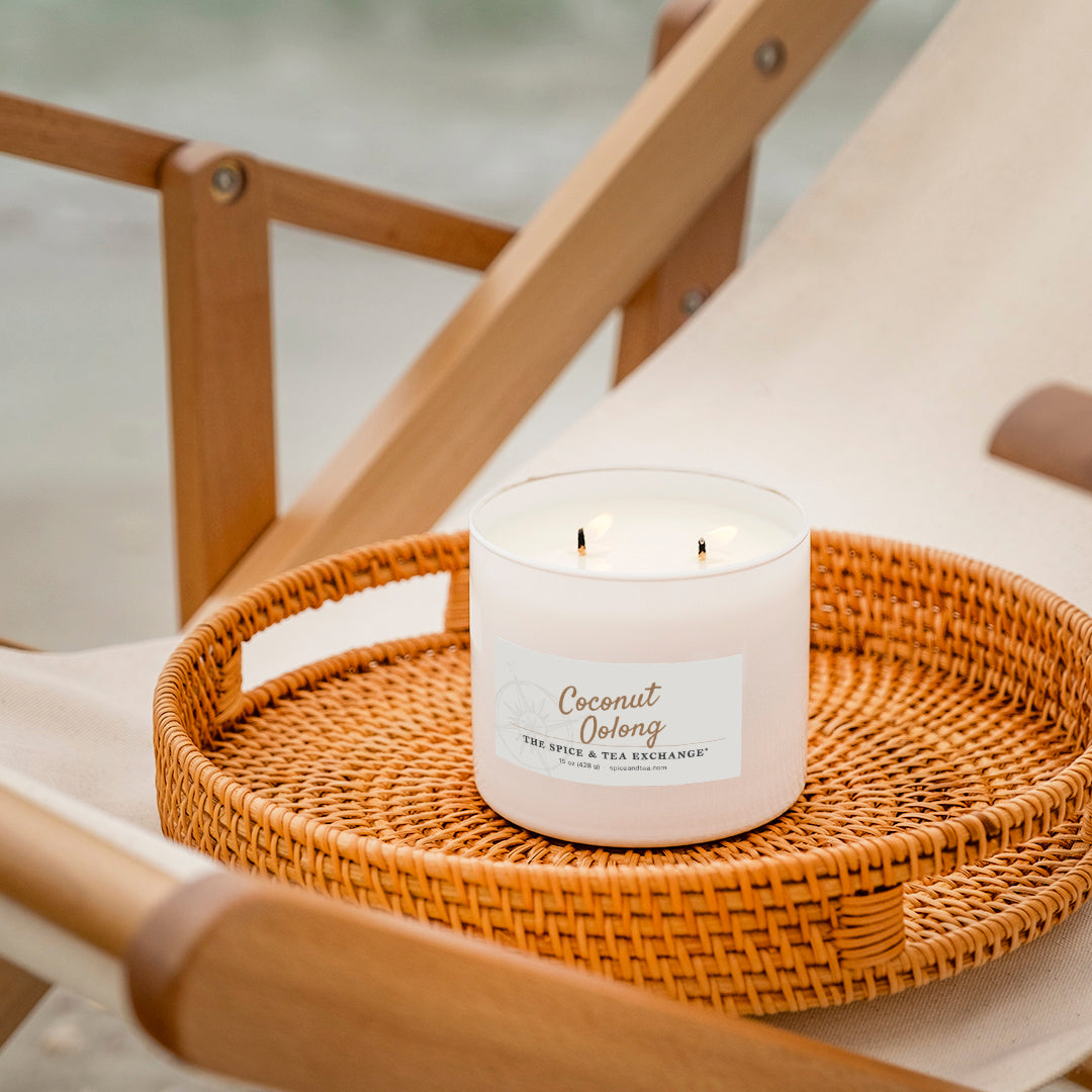 A white candle labeled 'Coconut Oolong' sits on a round woven tray placed on a wooden beach chair.