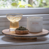 A glass of iced tea, a Coconut Oolong Candle made with essential coconut oils, and a wooden bowl of loose leaves sit on a round tray by the window. Hand poured in the USA, this candle brings quality and elegance to any space.
