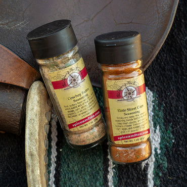 Two spice seasoning bottles labeled Spiceland's 'Cowboy ButterSeasoning' and 'Elote Street Corn Seasoning' resting on a rustic leather and woven textile background.