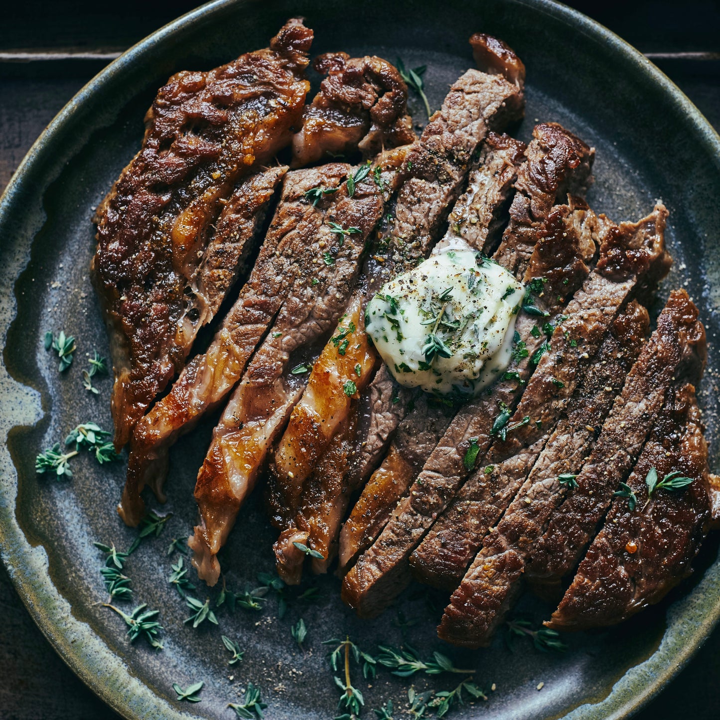 Sliced grilled steak on a dark ceramic plate with a dollop of butter topped with herbs, garnished with thyme.