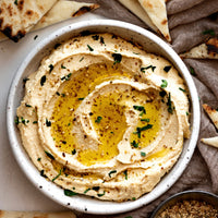 A top-down view of a white speckled bowl filled with creamy hummus, drizzled with olive oil and garnished with chopped parsley and herbs, surrounded by pieces of flatbread on a neutral fabric background.