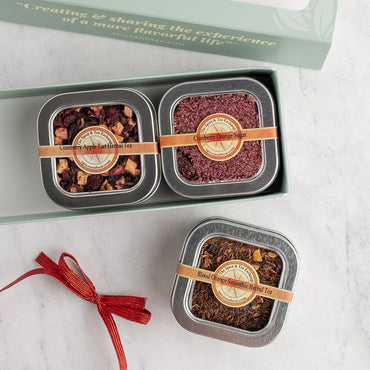 A boxed set of three square metal tea tins displayed on a light marble surface. The open mint-green gift box at the top shows two tins inside, and a third tin sits below.