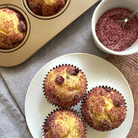 Three muffins (with cranberries in them topped with a red sugar) on a white plate, with two more in a metal muffin pan and a small bowl of red sugar nearby on a patterned gray cloth.
