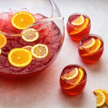 A glass pitcher filled with red iced beverage and sliced orange and lemon, with three matching small glasses arranged on a light surface.