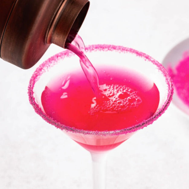 Bright pink liquid being poured from a dark metal pitcher into a martini glass with a pink sugar-rimmed edge, on a clean white background.