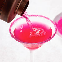 Bright pink liquid being poured from a dark metal pitcher into a martini glass with a pink sugar-rimmed edge, on a clean white background.