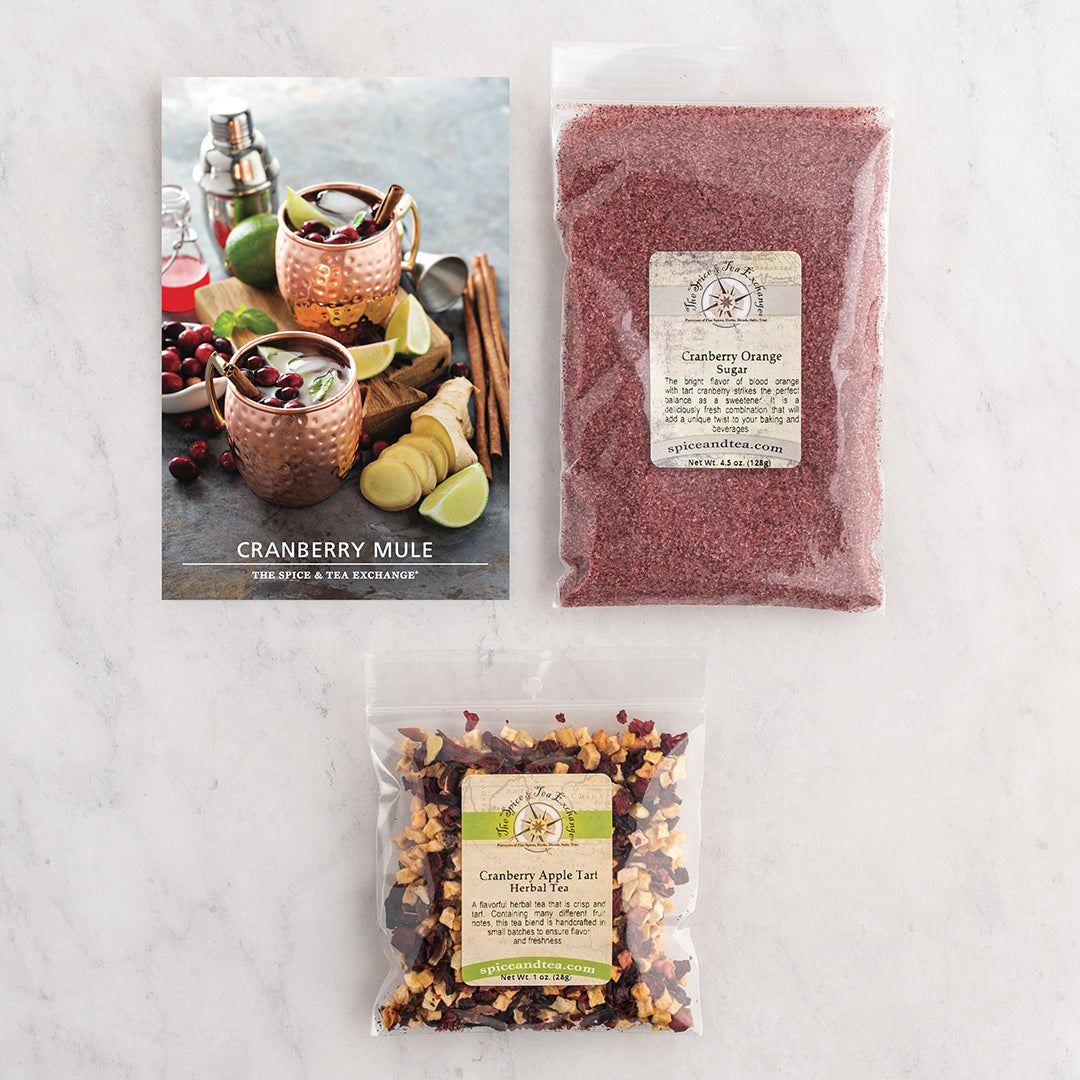 Three packaged food items arranged on a light marble surface: a printed recipe card for a cranberry mule, a clear bag of pinkish-red cranberry orange spice blend, and a clear bag of cranberry spice tea with mixed herbs and fruit.