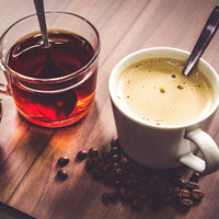 A white ceramic coffee mug filled with espresso and a clear glass mug filled with iced tea on a wooden table, with coffee beans scattered around them.